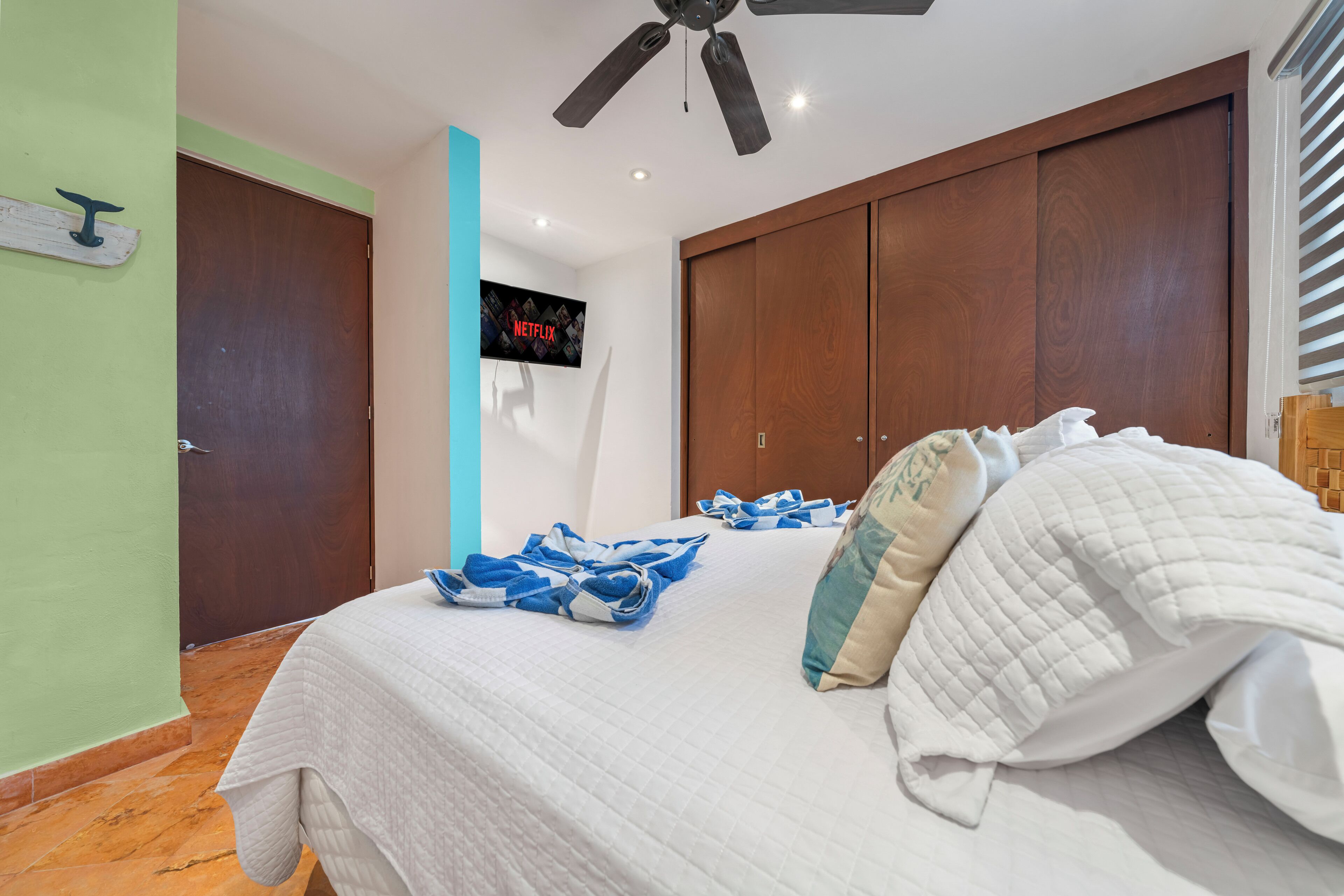 A bedroom with a bed, a ceiling fan, a TV displaying the Netflix logo, and a wooden wardrobe.