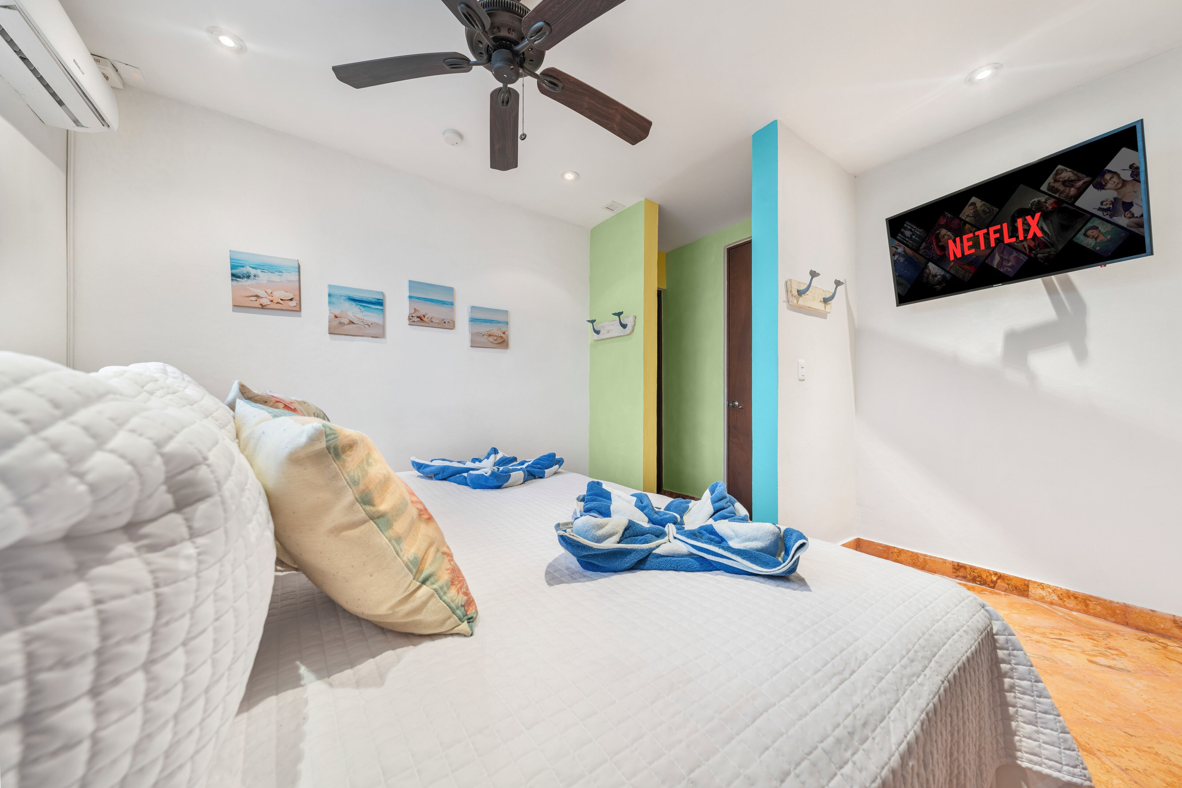 A hotel room with a bed, a ceiling fan, a wall-mounted TV displaying the Netflix logo, and a wall with three doors.