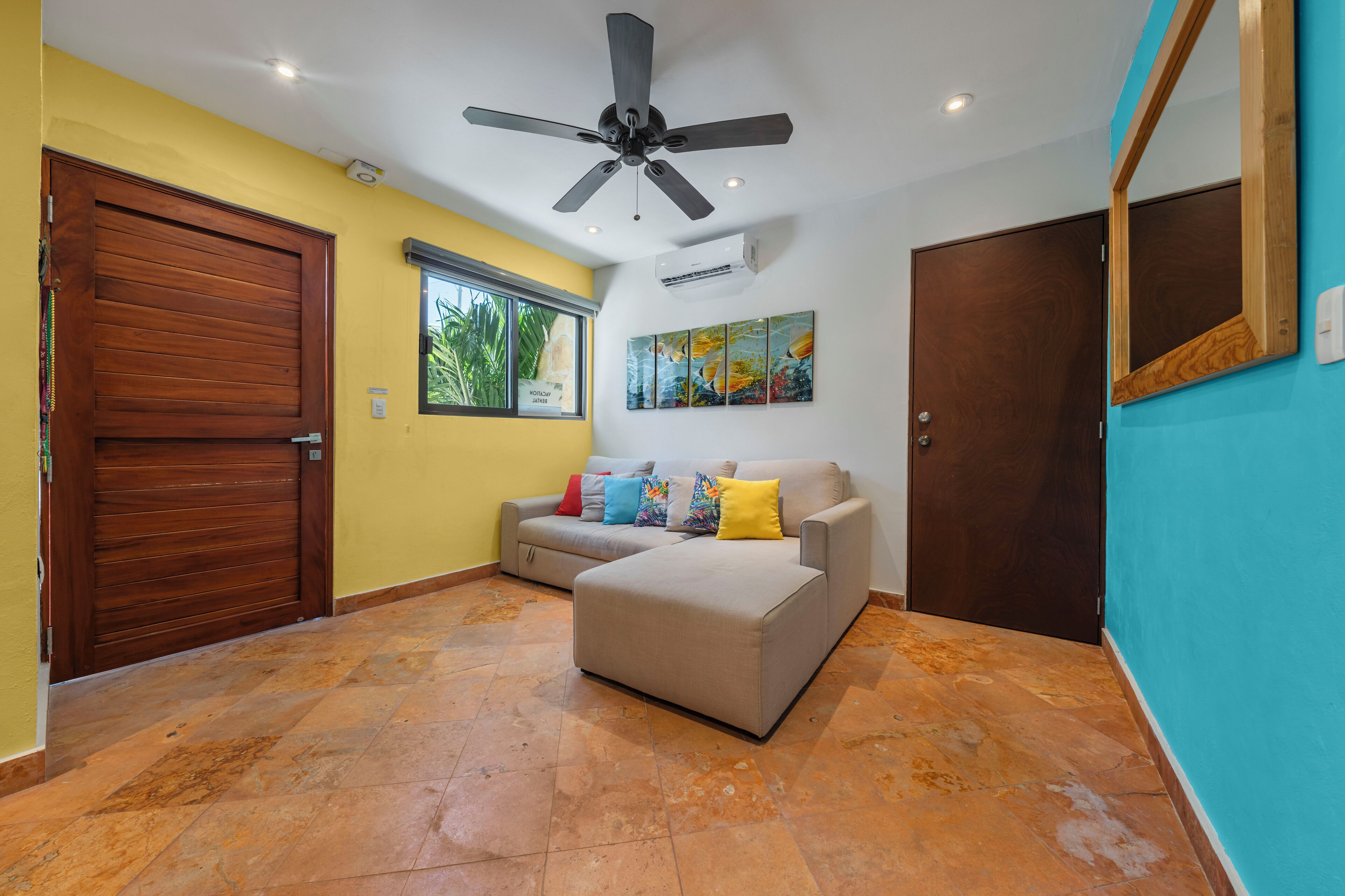 A room with a ceiling fan, a wooden door, a beige sofa with colorful cushions, a window, and a wall-mounted air conditioner.