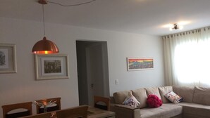 A living room with a beige sofa, a dining table with chairs, a pendant light, framed pictures on the wall, and a window with curtains.