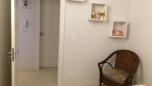 A room with a wicker chair, a bed with a floral bedspread, and three white shelves on the wall.