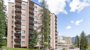 A multi-story residential building with balconies, surrounded by greenery and mountains in the background.