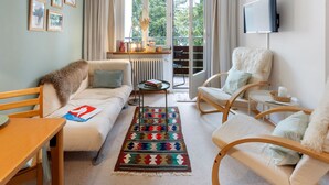 A cozy living room with a sofa, chairs, a coffee table, and a TV. There is a large window with curtains, a radiator, and a bookshelf with books and decorative items.