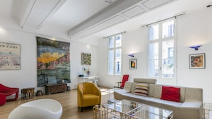 Un amplio salón con un sofá moderno, un sillón amarillo y una silla blanca. Grandes ventanales dejan entrar luz natural y se exhiben obras de arte en las paredes.