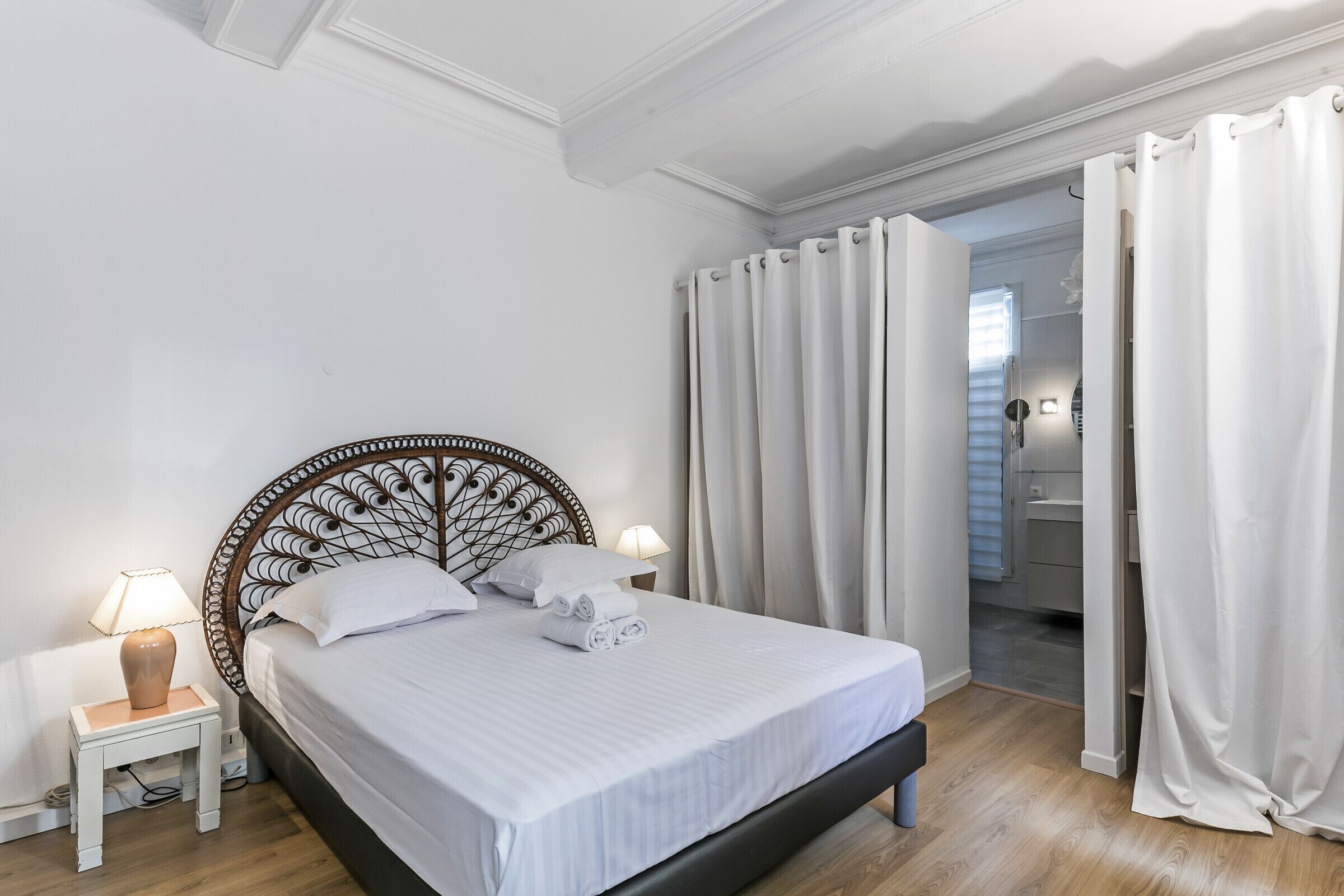 A neatly made bed with white linens and a headboard, a bedside lamp, and a view into a bathroom through a curtained doorway.