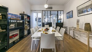 Un comedor moderno con una mesa blanca, sillas y una mesa de vidrio con un florero. Hay estantes con objetos decorativos y una pintura de un paisaje urbano en la pared.