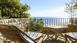 Une terrasse en bois avec deux chaises longues à rayures et une petite table, donnant sur un mur de pierre et offrant une vue sur la mer.