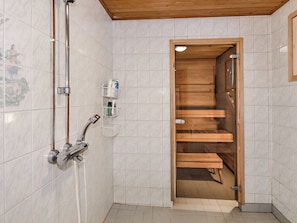 Un baño con paredes de azulejos blancos, un banco de madera, una ducha con accesorios cromados y un perchero con artículos de aseo.