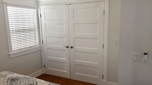 A bedroom with a double door, a window with blinds, a patterned bedspread, and a thermostat on the wall.