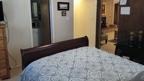 A bedroom with a bed, wooden dresser, and a door leading to a kitchen.