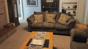 A living room with a brown sofa, patterned cushions, a wooden coffee table, and a bookshelf with decorative items.