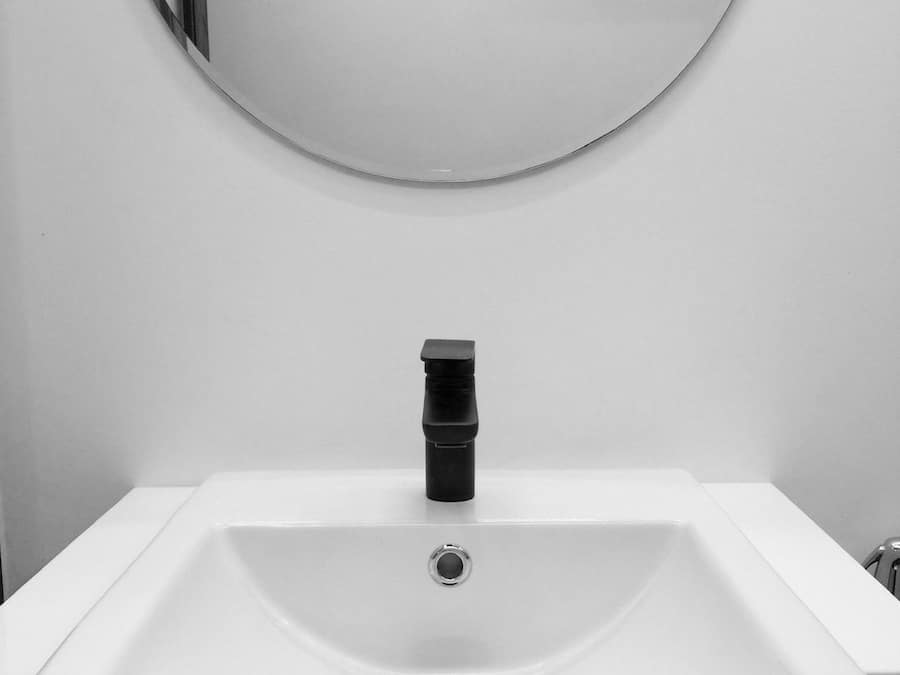 A white bathroom sink with a black faucet, a round mirror above it, and a towel rack to the right.