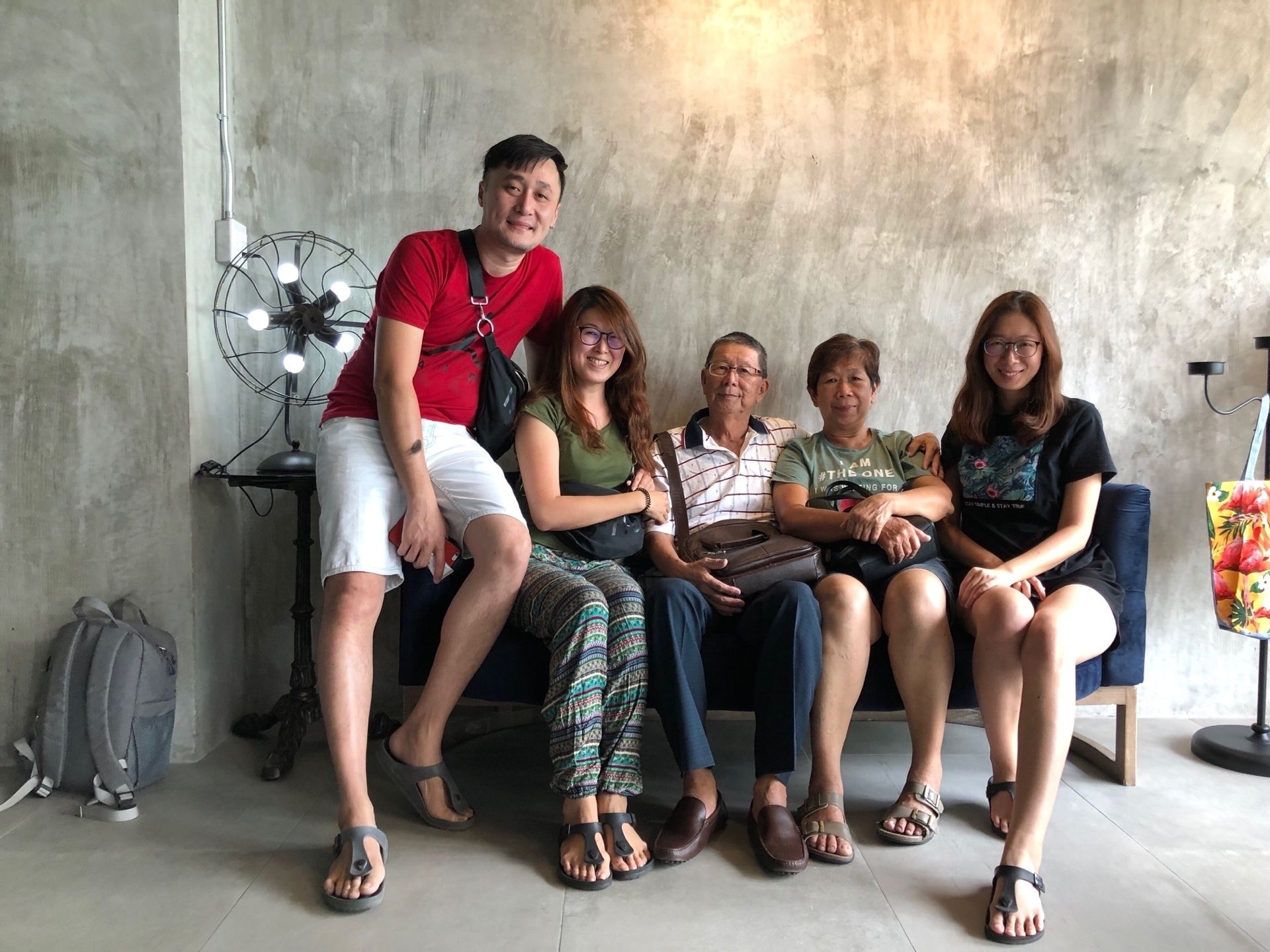 A group of people seated on a couch in a room with a concrete wall and a standing fan.