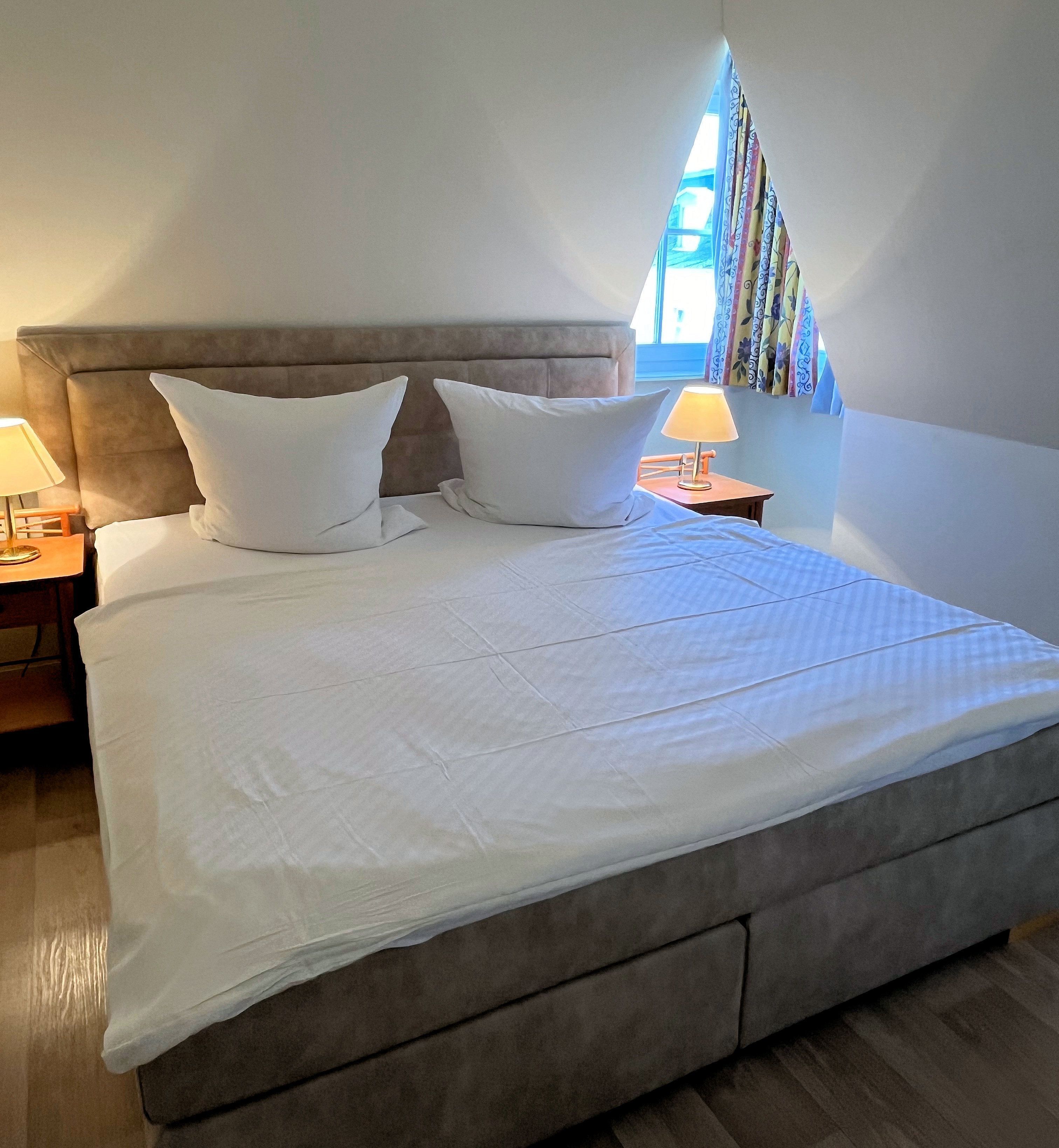 Una cama bien hecha con sábanas y almohadas blancas, cabecera de madera, mesita de noche con lámpara y ventana con cortinas.