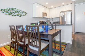 A modern kitchen with white cabinets, a stainless steel refrigerator, and a dining area with wooden chairs and a colorful rug.