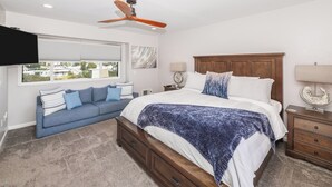 Un dormitorio con cama, cabecero de madera, ventilador de techo, ventana con persianas, sofá con cojines y mesita de noche con lámpara.
