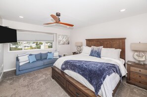 A bedroom with a bed, a wooden headboard, a ceiling fan, a window with blinds, a sofa with cushions, and a nightstand with a lamp.