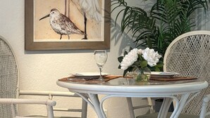 A dining area with a white metal table and chairs, a framed painting of birds on the wall, and a potted plant with white flowers. - A Perfect Condo Near The Beach - just renovated! (St Pete Beach)