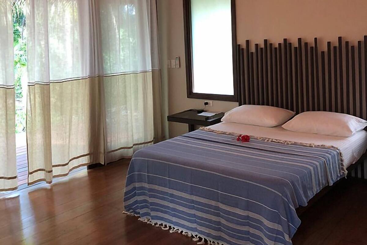 A bedroom with a bed, a nightstand, a window with curtains, and a wall with vertical slats.