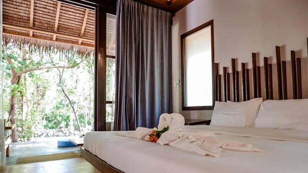 A hotel room with a large bed, a wooden headboard, a window with curtains, and a view of greenery outside. - Sadhana Siargao (General Luna)