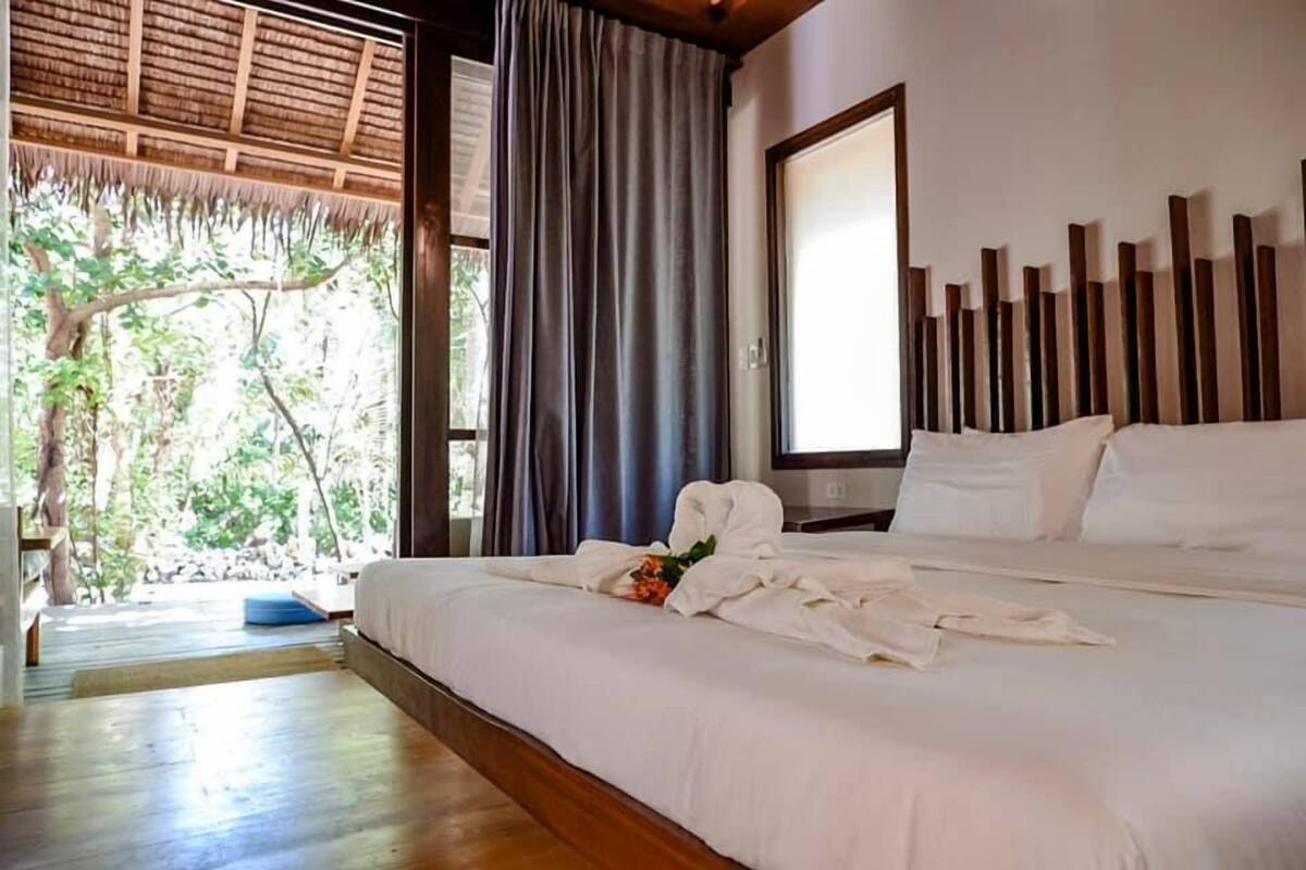 A hotel room with a large bed, a wooden headboard, a window with curtains, and a view of greenery outside.