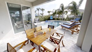 Une terrasse extérieure avec une table et des chaises en bois, un barbecue, des chaises longues près d’une piscine et des palmiers en arrière-plan.