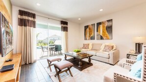 A modern living room with a sofa, coffee table, and two armchairs. There is a television on a wooden stand and a dining area visible through a glass door. - Encore Resort at Reunion 1855 (Kissimmee)