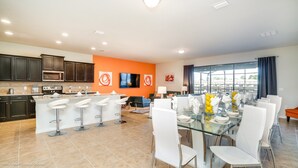 A modern kitchen with dark cabinets, a central island with bar stools, and a dining area with a glass table and white chairs. - Windsor at Westside 8817 (Kissimmee)