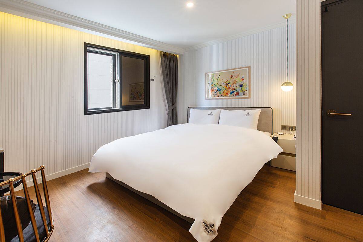 A bedroom with a large bed, a window with curtains, a bedside table, and a door.