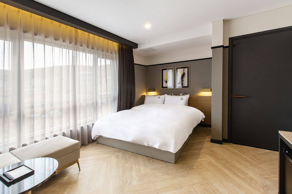 A modern hotel room with a large bed, a sitting area with a table, and a view through sheer curtains.