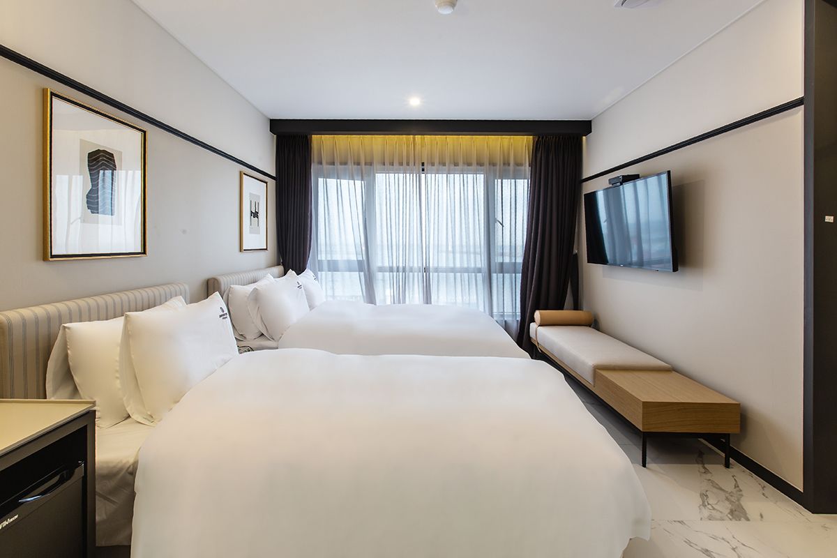A hotel room with two beds, a TV, a desk, and a large window with curtains.