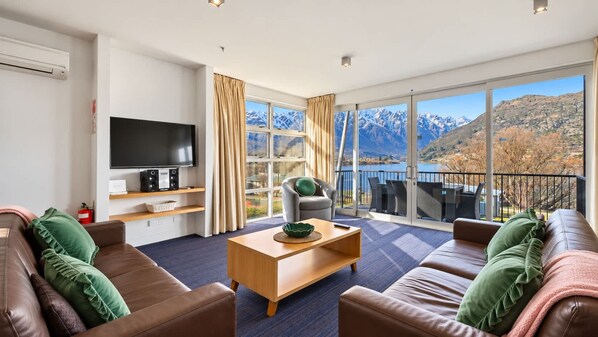 A modern living room with a flat-screen TV, leather sofas, a wooden coffee table, and a large window offering a mountain view.