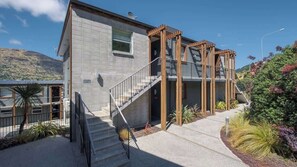 Une maison moderne à deux étages, avec une façade en béton, un escalier en bois et une pergola dotée d’une rambarde en métal.