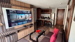 A modern living room with a flat-screen TV displaying a streaming service interface, a kitchen area with bar stools, and a cozy seating area with a sofa and a coffee table.