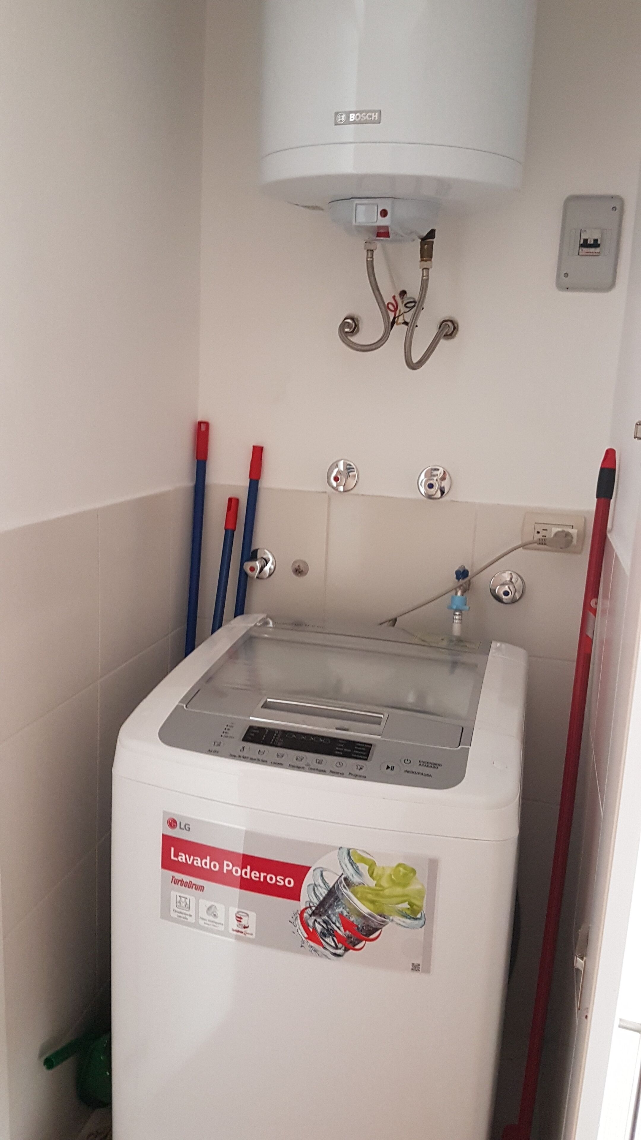 Màquina de rentar compacta i blanca amb panell de control i un dipòsit d'aigua, muntada a la paret amb una presa d'electricitat i una escoba vermella a prop.