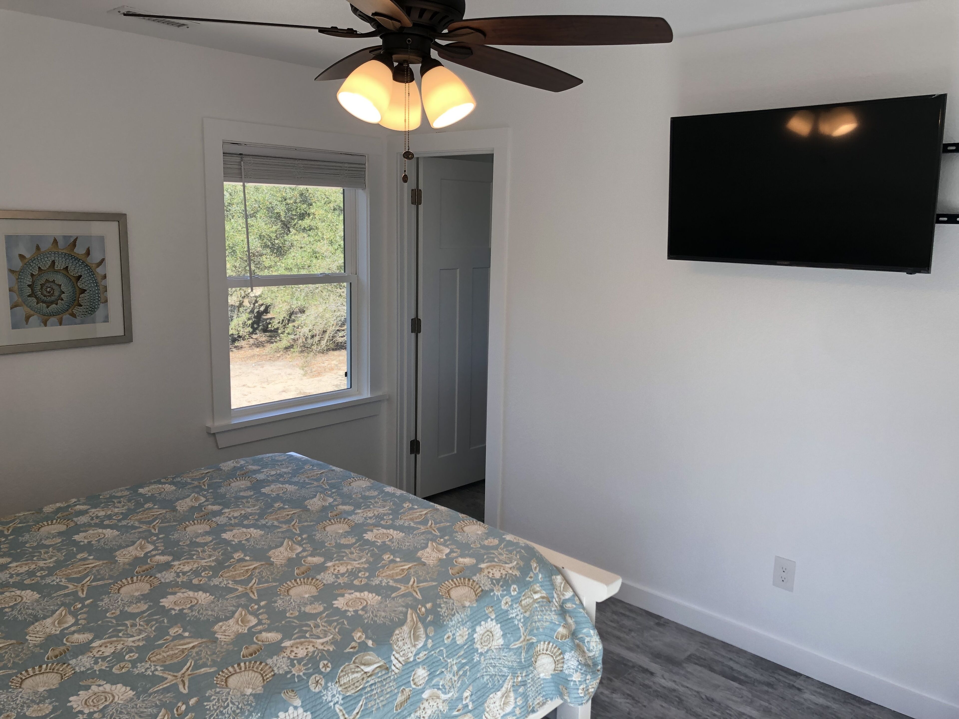 A bedroom with a bed, a ceiling fan, a window, a door, and a TV mounted on the wall.