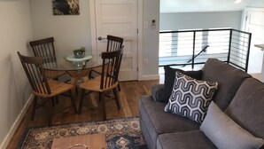 A dining area with a glass table and wooden chairs, a gray sofa with patterned pillows, and a view of a balcony with a railing.