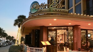 Das Außere eines Restaurants namens "Patio Bar & Grill" mit einem geschwungenen Eingangsbereich, Sitzplätzen im Freien und Palmen im Hintergrund.