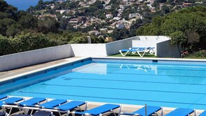 Una piscina amb gandules blaves i blanques, envoltada de vegetació i amb vista a un poble situat en un turó.