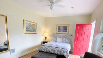 A bedroom with a bed, a ceiling fan, a red door, a black chair, and a bench.