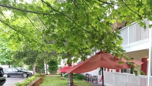 A well-maintained garden with a lawn, a table, and chairs under a red umbrella, surrounded by a white fence and a building with balconies. - The Caden Hotel (Vernonia)
