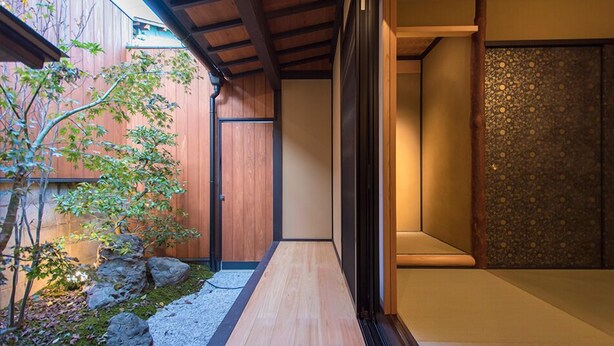 Un intérieur japonais traditionnel avec un plancher en bois, une pièce avec des tatamis et un petit jardin agrémenté de rochers et d’un arbre.
