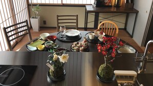 Un coin repas avec une table dressée pour deux personnes, où l’on sert des plats traditionnels japonais, agrémenté d’un vase de fleurs blanches et d’un bonsaï.