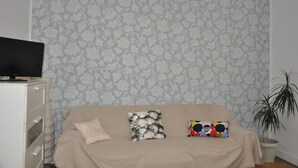 Een kamer met een beige bank, twee gekleurde kussens, een televisie op een witte kast en een pot met een plant.