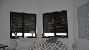 Een hotelkamer met een bed, twee ramen met zonwering, een stoel, een tafel en een lamp.