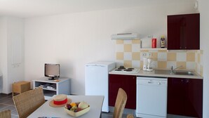 A compact kitchen with a dishwasher, a coffee maker, and a microwave. There is a dining table with chairs and a basket of fruit.