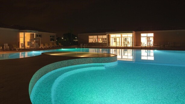 A nighttime swimming pool with illuminated edges, lounge chairs, and a building with open doors.