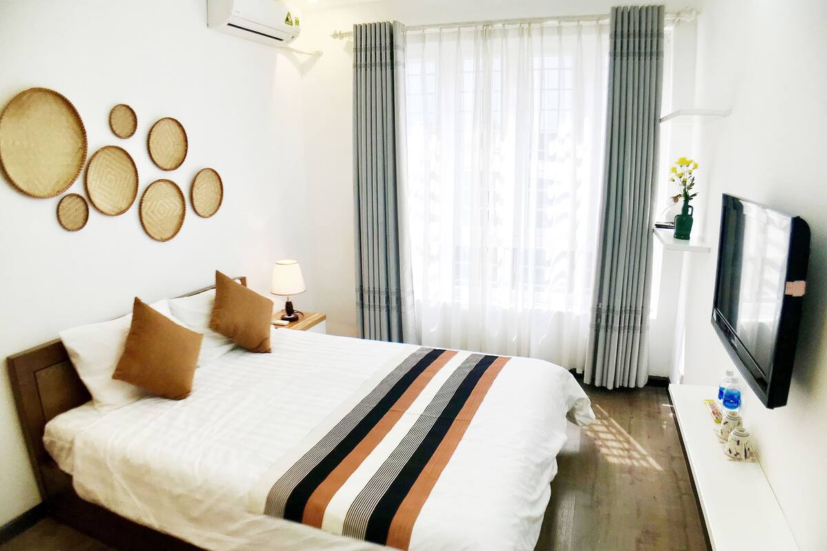 A hotel room with a bed, a TV, a bedside table with a lamp, a wall-mounted air conditioner, and a window with curtains.