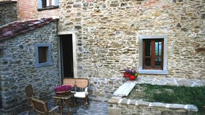 Un bâtiment en pierre doté d’un petit patio aménagé avec des chaises et une table en osier, une fenêtre avec un jardinier et une porte.