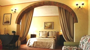 Une chambre avec une grande porte en arc de cercle, un lit recouvert d’une couvre-lit à motifs floraux, une table de chevet, une lampe et une chaise.